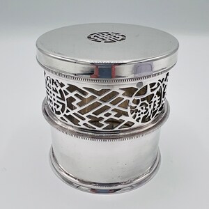 May include: Silver cylindrical box with a decorative, openwork design. The box has a lid with a raised, circular design.