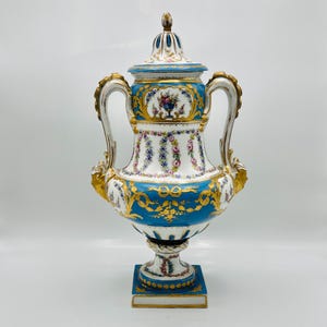 Puede incluir: Un jarrón decorativo de porcelana con tapa, en azul y blanco con detalles dorados. El jarrón tiene dos asas y está adornado con motivos florales y detalles ornamentados. Se apoya sobre una base cuadrada. Altura aproximada 40 cm.