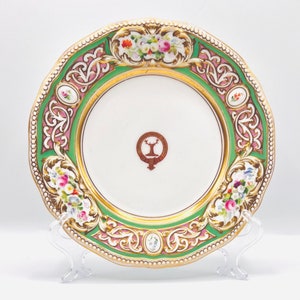 May include: A white porcelain plate with a green and pink floral design. The plate has a gold rim and a crest in the center.