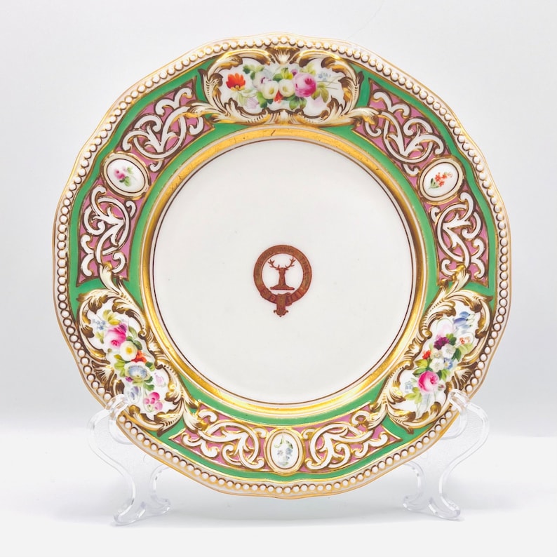 May include: A white porcelain plate with a green and pink floral design. The plate has a gold rim and a crest in the center.