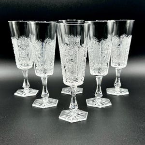 Puede incluir: Seis copas de champán de cristal transparente con intrincados diseños de estrellas y geométricos. Cada copa presenta una copa alta y delgada, un tallo delicado y una base hexagonal. Las copas están sobre una superficie oscura.