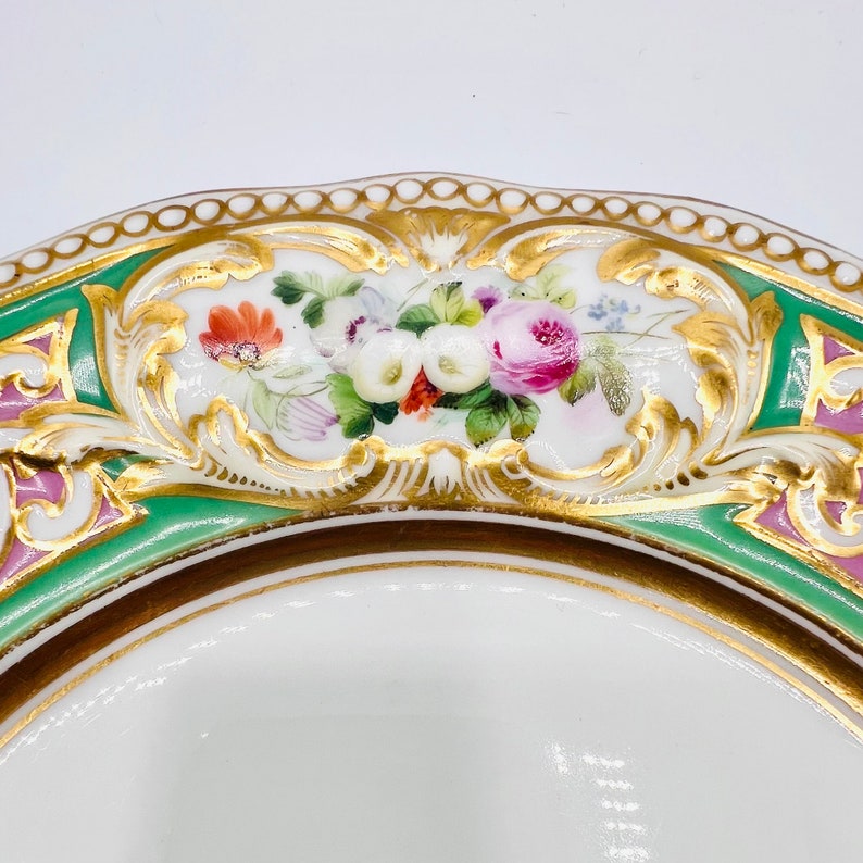 May include: A white porcelain plate with a green and pink border and gold trim. The center of the plate features a hand-painted floral design with pink, white, and red flowers.