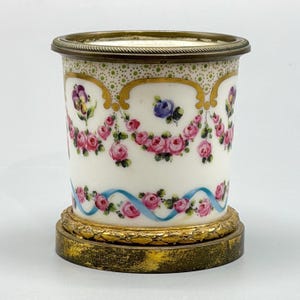 Puede incluir: Un recipiente cilíndrico de porcelana con fondo blanco, pintado a mano con guirnaldas de rosas rosadas, cintas azules y pensamientos morados. El borde superior y la base son de latón dorado con detalles decorativos texturizados.