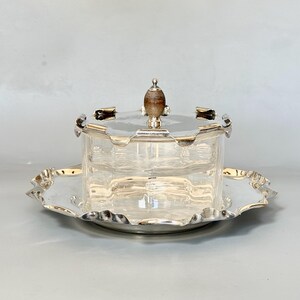 Puede incluir: Una mantequillera de cristal transparente con base y tapa plateadas. La tapa presenta un borde festoneado y un pomo central de madera. La base es una bandeja plateada a juego con un borde festoneado decorativo, ofreciendo un acabado pulido.