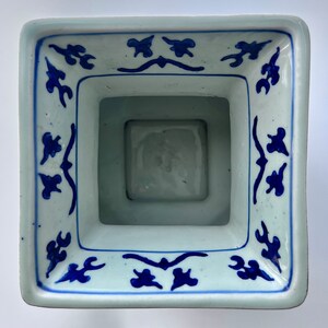 Puede incluir: Un macetero cuadrado de cer&aacute;mica blanca con un dise&ntilde;o floral azul y blanco. El macetero tiene una abertura cuadrada en la parte superior y una base cuadrada.