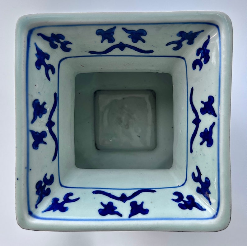 Puede incluir: Un macetero cuadrado de cer&aacute;mica blanca con un dise&ntilde;o floral azul y blanco. El macetero tiene una abertura cuadrada en la parte superior y una base cuadrada.