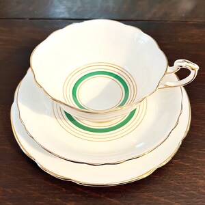 Puede incluir: Un juego de taza y platillo de t&eacute; blanco con un dise&ntilde;o de c&iacute;rculos conc&eacute;ntricos verde y dorado. El juego tiene un borde delicado y festoneado.
