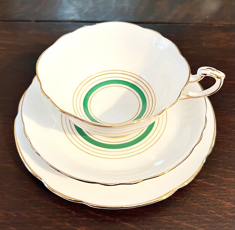 Puede incluir: Un juego de taza y platillo de t&eacute; blanco con un dise&ntilde;o de c&iacute;rculos conc&eacute;ntricos verde y dorado. El juego tiene un borde delicado y festoneado.