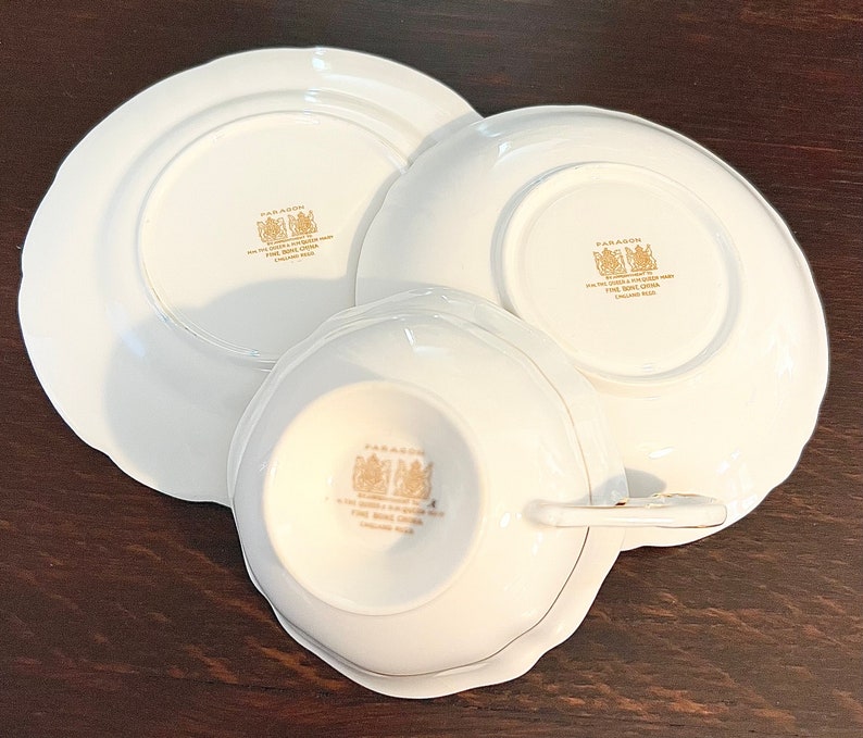 Puede incluir: Un juego de tres tazas de t&eacute; y platillos de porcelana blanca con ribetes dorados y las palabras "Paragon Fine Bone China England" en la parte inferior.