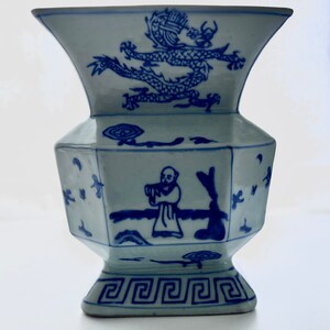 Puede incluir: Un jarr&oacute;n de porcelana azul y blanco con un dise&ntilde;o de drag&oacute;n en la parte superior y una figura tocando la flauta en el lateral. El jarr&oacute;n tiene una base cuadrada con un borde decorativo.
