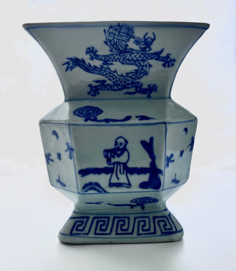 Puede incluir: Un jarr&oacute;n de porcelana azul y blanco con un dise&ntilde;o de drag&oacute;n en la parte superior y una figura tocando la flauta en el lateral. El jarr&oacute;n tiene una base cuadrada con un borde decorativo.