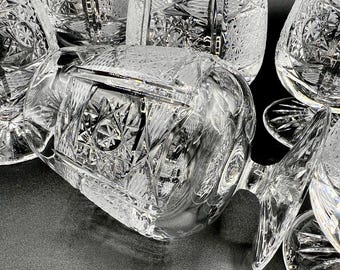 May include: A selection of clear crystal glassware with detailed cut patterns. The image shows various glasses, including wine glasses and tumblers, with intricate geometric designs. The glasses are arranged on a dark surface, emphasising their clarity and craftsmanship.