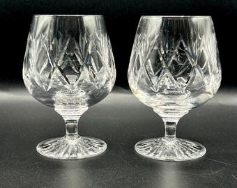 May include: Two clear crystal brandy glasses with diamond-cut patterns. The glasses have a rounded bowl shape, a short stem, and a wide, circular base. The crystal reflects light. The background is dark.
