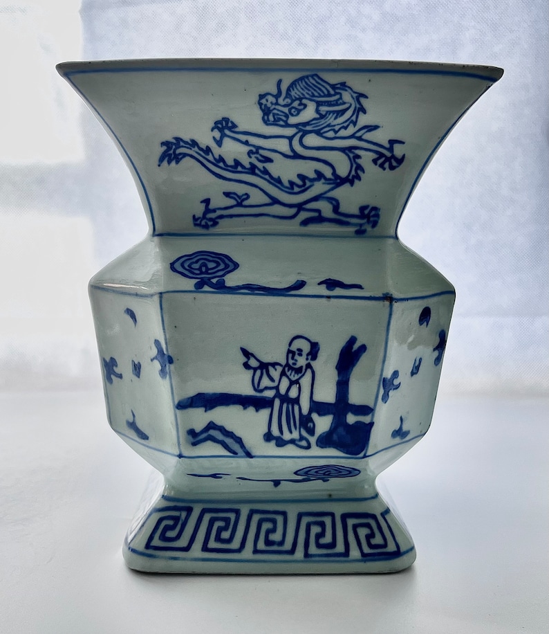 Puede incluir: Un jarr&oacute;n de porcelana azul y blanco con un dise&ntilde;o de drag&oacute;n y una figura. El jarr&oacute;n tiene una base cuadrada y una boca ancha.