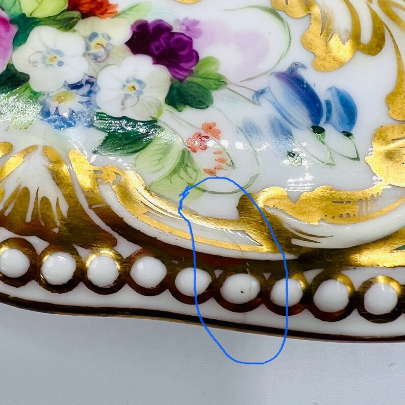 May include: A white porcelain trinket box with a floral design and gold accents. The box has a heart shape and a beaded trim around the edge.