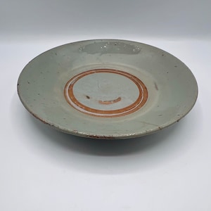 Peut inclure: Une assiette en céramique vert clair avec un anneau brun autour du centre. L'assiette a une texture légèrement rugueuse et est légèrement ébréchée sur les bords.