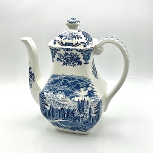 Puede incluir: Una tetera de cerámica blanca con un diseño de paisaje azul y blanco. La tetera presenta un pico curvo, un asa grande y una tapa con un estampado floral. La escena del paisaje representa un castillo y árboles.