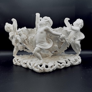 May include: A white porcelain centerpiece bowl featuring three sculpted cherub figures standing around a central fluted basin. The base is decorated with intricate openwork lattice patterns and sculpted thistle floral motifs throughout.