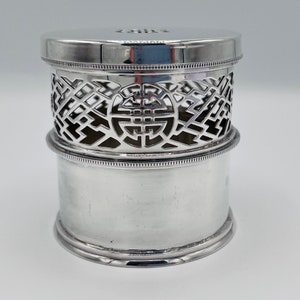 Pode incluir: Caixa cilíndrica de prata com uma tampa de trabalho aberto decorativa que apresenta um design de nó chinês. A tampa é ligeiramente elevada e tem uma superfície lisa e polida.