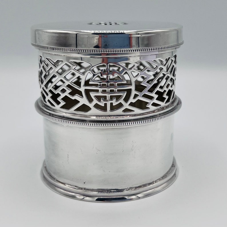 May include: Silver cylindrical box with a decorative openwork lid featuring a Chinese knot design. The lid is slightly raised and has a smooth, polished surface.