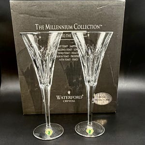 Peut inclure: Deux flûtes à champagne en cristal transparent avec des motifs taillés verticaux sur les bols, placées devant une boîte gris foncé étiquetée The Millennium Collection. La boîte présente du texte listant cinq toasts et le logo Waterford Crystal.