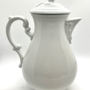 Puede incluir: Cafetera de porcelana blanca con tapa y asa adornada. La cafetera tiene un cuerpo redondeado y un pico con un diseño decorativo.