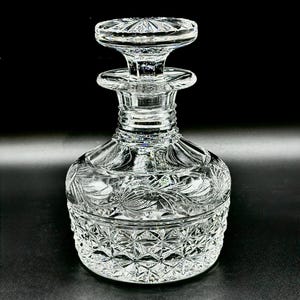 Puede incluir: Un decantador de vidrio transparente con una base ancha y redondeada con un patrón de corte de diamante y un cuello adornado con motivos florales grabados. El decantador incluye un tapón de vidrio a juego con un diseño de estrella y borde.