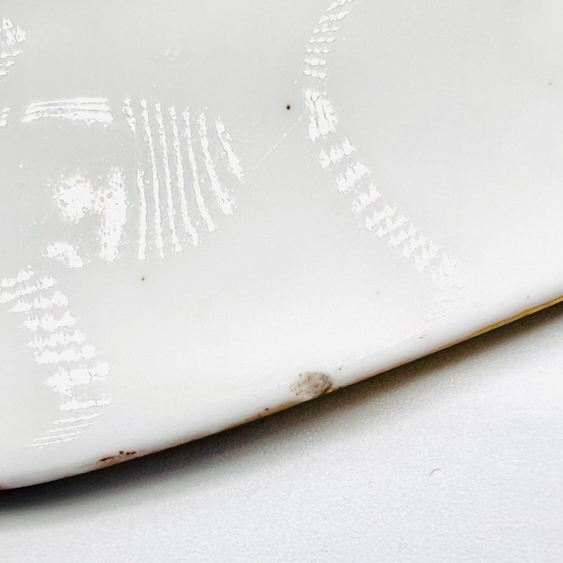 May include: White ceramic plate with a decorative design of white lines and dots. The plate has a slightly chipped edge.