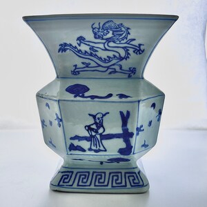 Puede incluir: Un jarr&oacute;n de porcelana azul y blanco con forma geom&eacute;trica y dise&ntilde;os intrincados. El jarr&oacute;n presenta un drag&oacute;n y una figura en un &aacute;rbol, todo pintado en azul sobre fondo blanco.