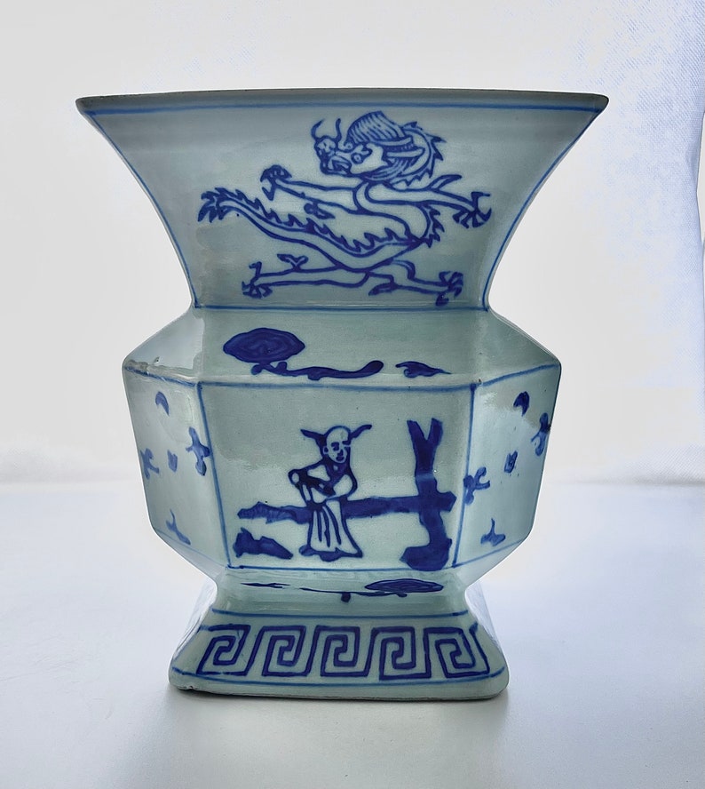 Puede incluir: Un jarr&oacute;n de porcelana azul y blanco con forma geom&eacute;trica y dise&ntilde;os intrincados. El jarr&oacute;n presenta un drag&oacute;n y una figura en un &aacute;rbol, todo pintado en azul sobre fondo blanco.