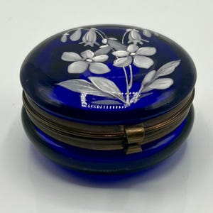 Peut inclure: Une boîte à bijoux ovale bleu cobalt avec un couvercle à charnière, ornée d'un motif floral blanc. La boîte a un bord et un fermoir en métal de couleur bronze. La boîte mesure environ 7 cm de long.