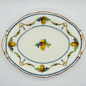 Puede incluir: Una fuente ovalada para servir con fondo blanco y borde azul y dorado. La fuente está decorada con un patrón floral de frutas y flores en tonos rojos, amarillos y verdes.