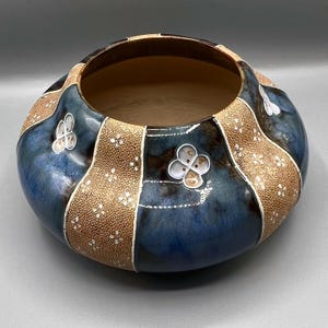 Op de afbeelding: Een decoratieve keramische kom met een brede, afgeronde vorm. De kom heeft een combinatie van blauwe, bruine en beige kleuren. De blauwe delen zijn glanzend, terwijl de beige delen een textuur met stippen hebben. Er zijn ook witte bloemmotieven aanwezig.