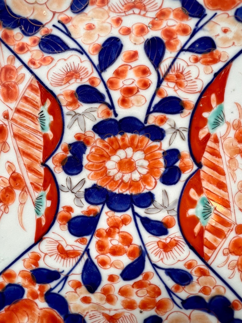 K&ouml;nnte beinhalten: Ein wei&szlig;er Keramikteller mit einem blauen und roten Blumenmuster. Die Blumen sind stilisiert und haben ein traditionelles japanisches Design. Der Teller ist mit goldenen Akzenten verziert.