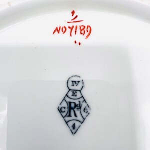 May include: Close-up of a white ceramic plate with a black and white diamond-shaped mark with the letters "CR" and the numbers "4" and "6" inside. The plate also has a red handwritten mark that says "NOT 189".
