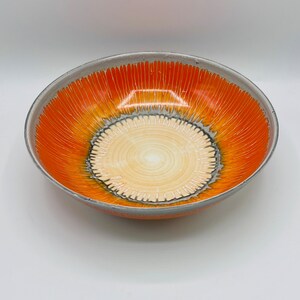 Könnte beinhalten: Eine große orangefarbene und graue Keramikschüssel mit einem konzentrischen Kreisdesign. Die Schüssel hat eine weiße Mitte mit einem grauen Rand und orangefarbene Streifen, die nach außen strahlen.