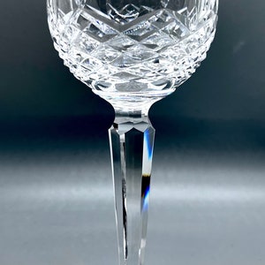 Può includere: Un bicchiere da vino in cristallo trasparente con una coppa rotonda e un gambo lungo e sottile. La coppa presenta un motivo tagliato e il gambo è sfaccettato. Anche la base del bicchiere è tagliata.