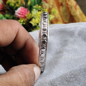 May include: A silver-colored bangle bracelet with a repeating pattern of small circles and vertical lines. The bracelet is held in a hand, showcasing its design and texture. The jewelry is likely made of metal.