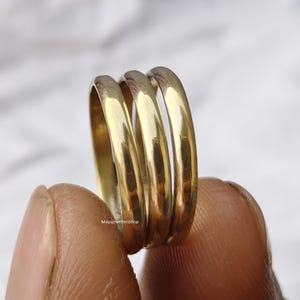 Op de afbeelding: Close-up van drie goudkleurige ringen die op elkaar zijn gestapeld. De ringen hebben een glad, glanzend oppervlak en een afgerond ontwerp. De ringen worden tussen twee vingers gehouden. De achtergrond is een vage, lichtgekleurde ondergrond.