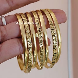 Peut inclure: Un ensemble de six bracelets rigides de couleur dorée présentant une alternance de surfaces lisses et polies et de gravures florales complexes. Le texte MayaJewellersShop est visible sur l'un des bracelets. Ces bijoux circulaires sont alignés.