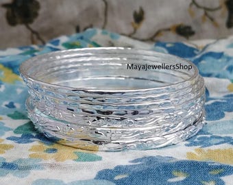 Könnte beinhalten: Ein Stapel silberner Armreifen mit strukturiertem, gehämmertem Design. Die Armreifen haben ein Blumenmuster und werden auf einem blau-gelben Stoff mit Blumenmuster präsentiert. Der Shop-Name "MayajewellersShop" ist sichtbar.