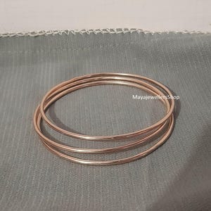 Puede incluir: Tres pulseras de oro rosa apiladas una encima de la otra sobre un fondo de tela gris. Las pulseras tienen todas el mismo ancho y un acabado liso y pulido.