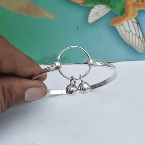 May include: A silver bracelet with a circular design and two small, dangling bell charms. The bracelet is held by a hand against a blurred background. The bracelet is made of a shiny, reflective metal.