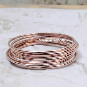 May include: A set of 10 rose gold bangles, stacked and arranged in a circle. The bangles are thin and delicate, with a smooth, polished finish.