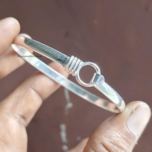 Puede incluir: Una pulsera de plata con cierre de gancho y ojo. La pulsera tiene un diseño simple y elegante con una banda redondeada y un cierre circular. El gancho está envuelto con alambre para mayor detalle. La pulsera se sostiene en una mano.