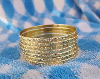 Könnte beinhalten: Ein Stapel goldfarbener Armreifen mit aufwendigen geätzten Designs. Die Armreifen liegen auf einer blau-weiß karierten Oberfläche. Der Schmuck stammt von MayajewellersShop.