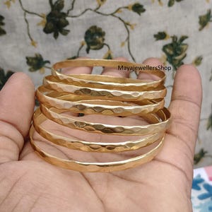 May include: A stack of six gold-colored bangles with a hammered texture. The bangles are stacked in a hand, with the text "MayajewellersShop" visible in the background.