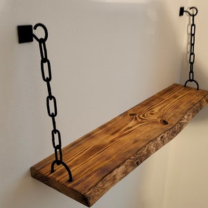 May include: A wooden shelf with a live edge, hanging from the ceiling with black chains. The shelf is stained a dark brown color and has a natural wood grain.