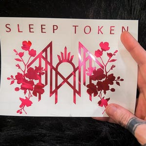 May include: A rectangular white decal featuring the text SLEEP TOKEN in red capital letters at the top. Below the text is a metallic red graphic design consisting of a central geometric emblem flanked by symmetrical floral branches on both sides.