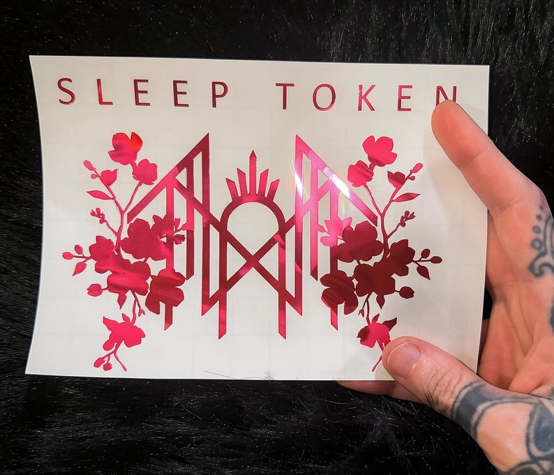 May include: A rectangular white decal featuring the text SLEEP TOKEN in red capital letters at the top. Below the text is a metallic red graphic design consisting of a central geometric emblem flanked by symmetrical floral branches on both sides.